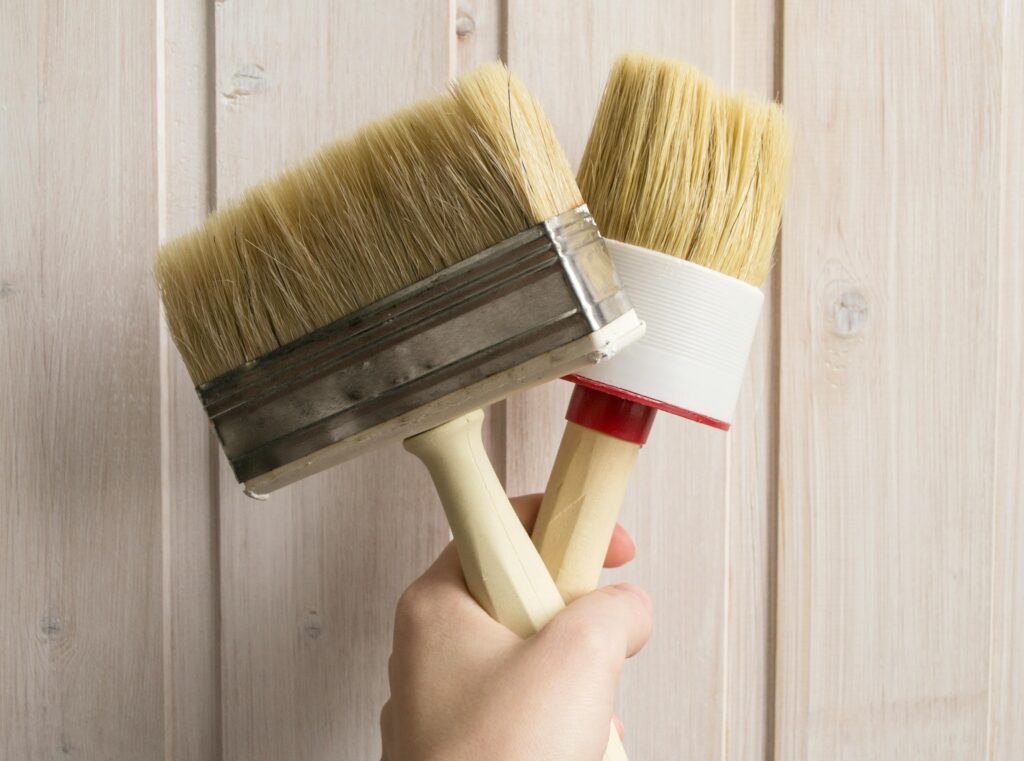 Close-up of a hand holding two large paintbrushes against a light wooden backdrop, ideal for DIY themes.
