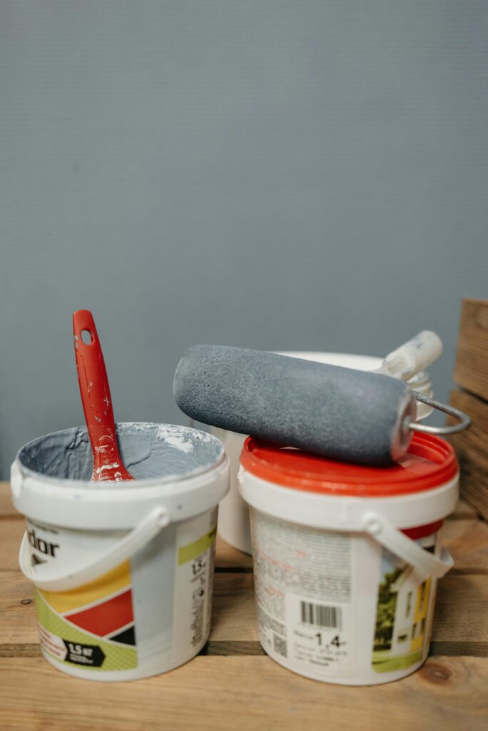 Close-up of paint buckets with brushes and roller on wooden surface for decorating projects.